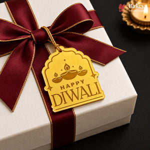 A gold-colored Diwali greeting tag with 'HAPPY DIwali' and diya illustrations, hanging from a dark red satin ribbon tied around a white gift box. A lit diya candle is visible in the background on a dark surface.