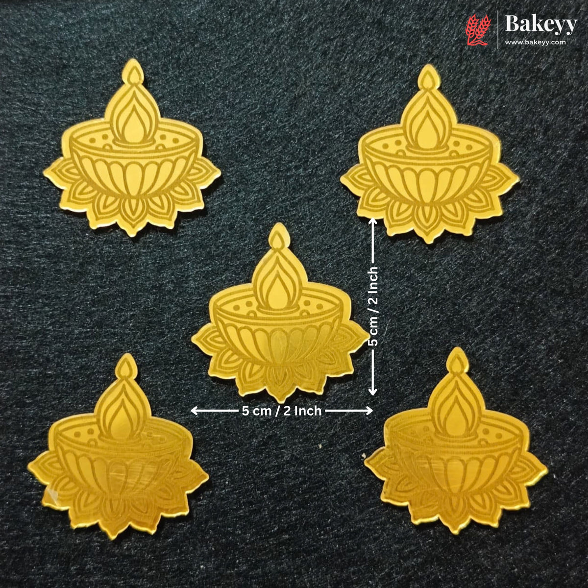 Five gold-colored metal oil lamp molds arranged on a dark textured background, with dimension arrows and text indicating a size of 5 cm by 5 cm (2 inches) around the central piece. A Bakeyy logo is visible in the top right corner.