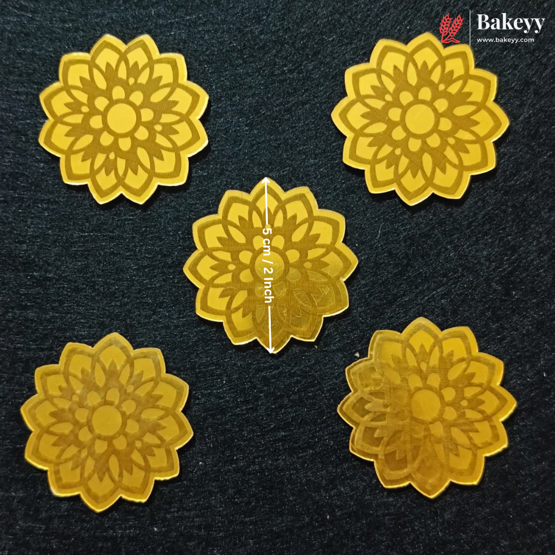 Five golden-yellow flower-shaped sugar paste or clay embellishments arranged on a dark textured background, with a central piece showing a vertical measurement marker of 5 cm / 2 inch. A Bakeyy logo is visible in the top right corner.