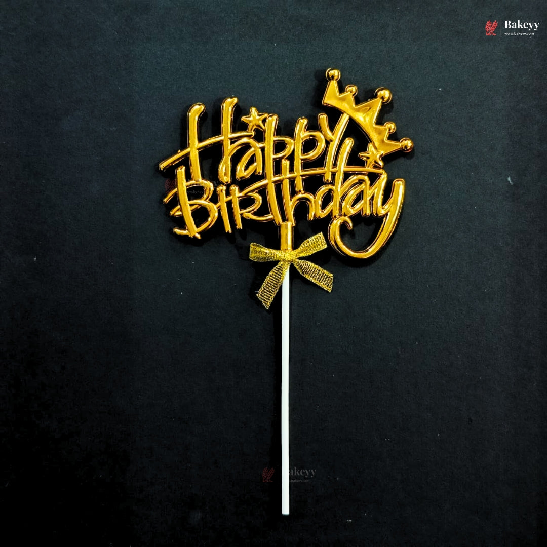 A gold metallic 'Happy Birthday' cake topper with a crown and bow design, mounted on a white stick, set against a dark black background.
