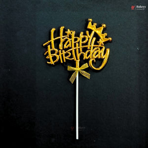 A gold metallic 'Happy Birthday' cake topper with a crown and bow design, mounted on a white stick, set against a dark black background.