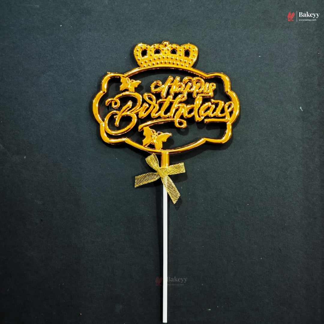 Gold-colored 'Happy Birthday' cake topper with a crown and bow on a stick, set against a dark textured background.