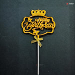 Gold-colored 'Happy Birthday' cake topper with a crown and bow on a stick, set against a dark textured background.