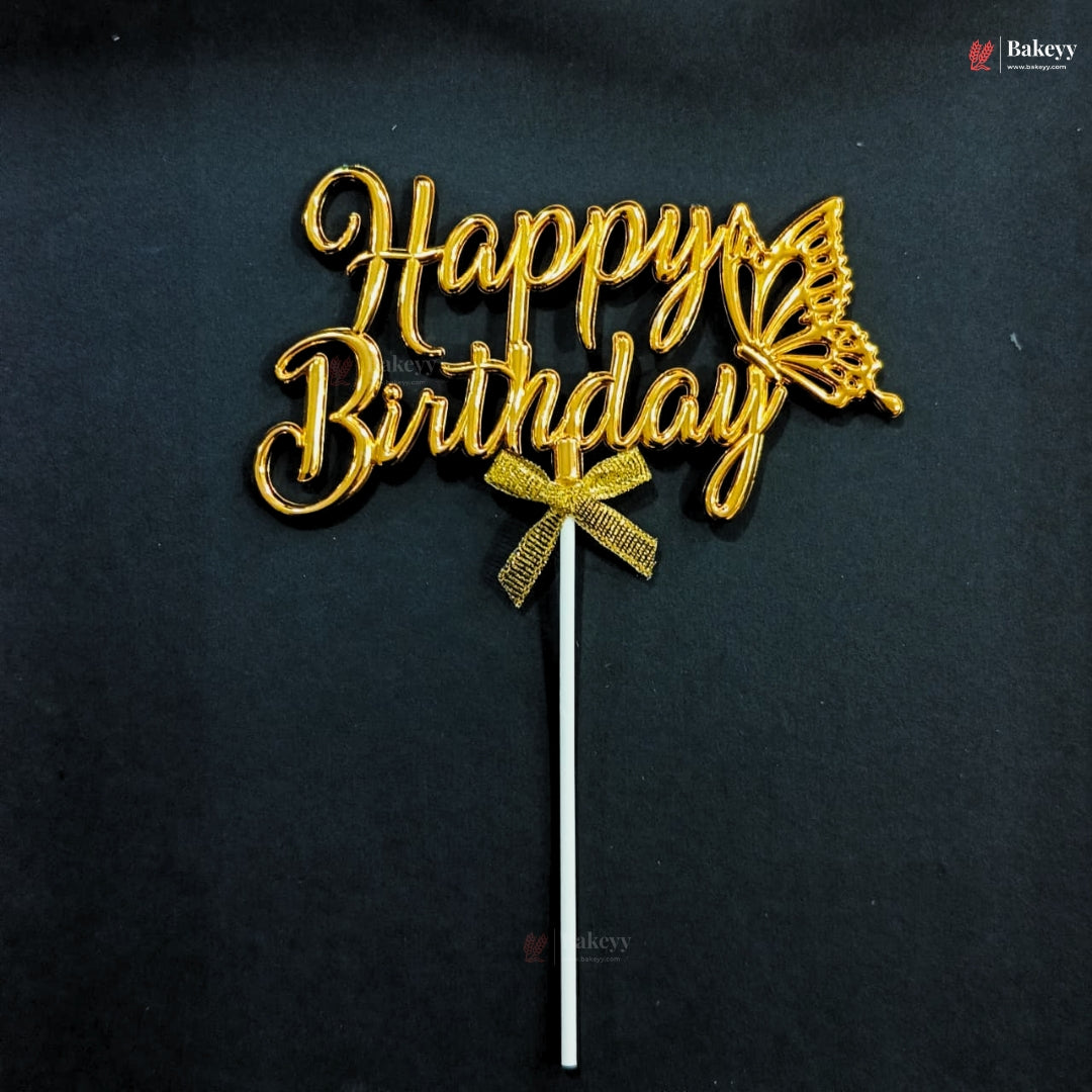 Gold metallic 'Happy Birthday' cake topper with a butterfly design and bow, mounted on a white stick, photographed against a dark background.