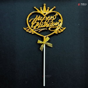 A gold-colored metal cake topper with a heart-shaped design, a crown on top, 'Happy Birthday' script, and a bow on a stick, set against a dark background.