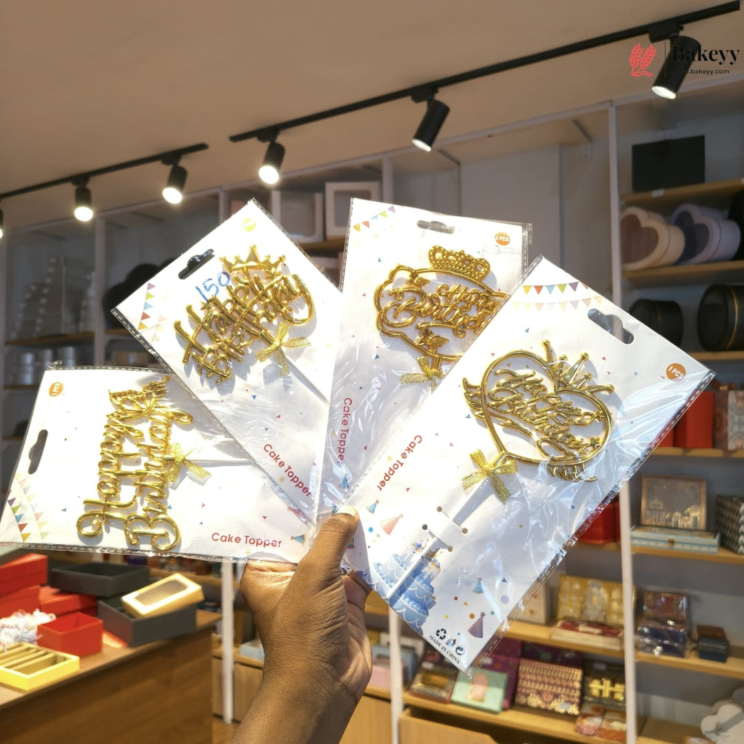 A hand holds four packaged gold cake toppers shaped like letters and a crown in a retail store. The packages are on white backing cards with confetti graphics, and store shelves with items are visible in the background under track lighting.