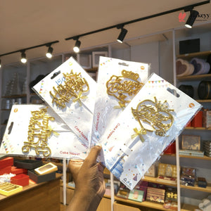 A hand holds four packaged gold cake toppers shaped like letters and a crown in a retail store. The packages are on white backing cards with confetti graphics, and store shelves with items are visible in the background under track lighting.