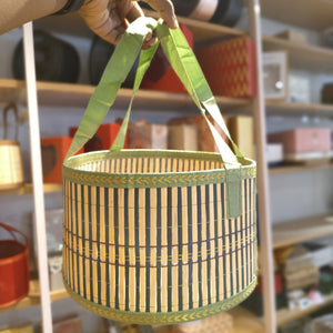 A hand holds a round woven basket with green fabric handles and a striped bamboo body, set against a blurred shop background.