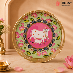 A decorative round serving tray with a gold-plated rim, featuring a pink and green floral pattern and a central illustration of a white cow and calf on a pink background. The tray is displayed on a marble surface with a warm beige background, lotus flowers, and a small brass lamp.