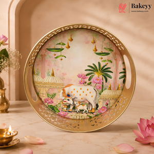 Round gold-plated serving tray with cutout handles, featuring a hand-painted traditional Indian illustration of a cow and calf under hanging lamps, displayed on a marble surface with decorative flowers and a lit brass lamp.