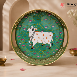 A decorative round serving tray with a gold-toned rim and handles, featuring a turquoise green center printed with a white cow, pink lotus flowers, and a geometric lattice pattern. The tray is styled on a light marble surface with a lit diya, scattered pink petals, and a warm beige background with arches.