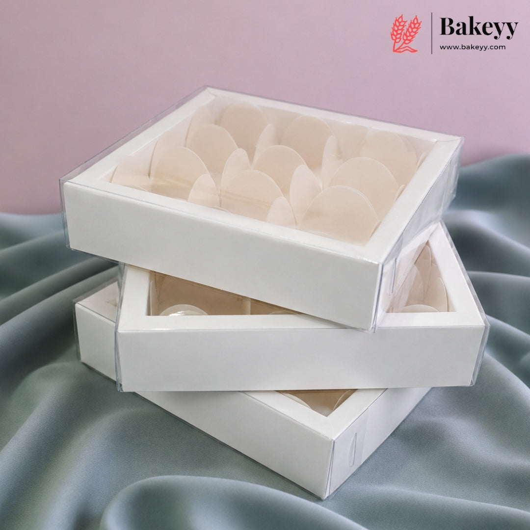Three white bakery display boxes with clear lids, stacked on draped satin fabric against a soft pink background. The top box contains pale cream-colored pastry inserts.