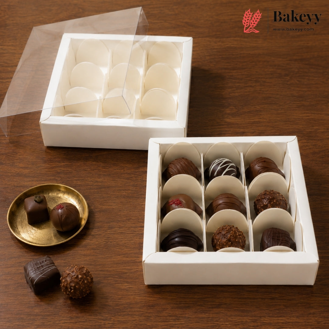 A white square chocolate box with a clear lid, shown open with multiple compartments holding assorted chocolates. A second empty compartment section of the same box is shown behind it, and two chocolates rest on a small gold plate beside the main box on a wooden surface.