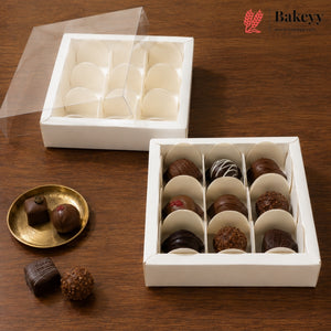 A white square chocolate box with a clear lid, shown open with multiple compartments holding assorted chocolates. A second empty compartment section of the same box is shown behind it, and two chocolates rest on a small gold plate beside the main box on a wooden surface.