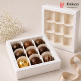 A white 12-compartment chocolate box shown open with assorted milk and dark chocolates inside, alongside the closed exterior of the same box. The scene is styled on a light beige surface with small white flowers and a beige ribbon.