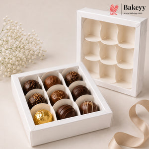 A white 12-compartment chocolate box shown open with assorted milk and dark chocolates inside, alongside the closed exterior of the same box. The scene is styled on a light beige surface with small white flowers and a beige ribbon.