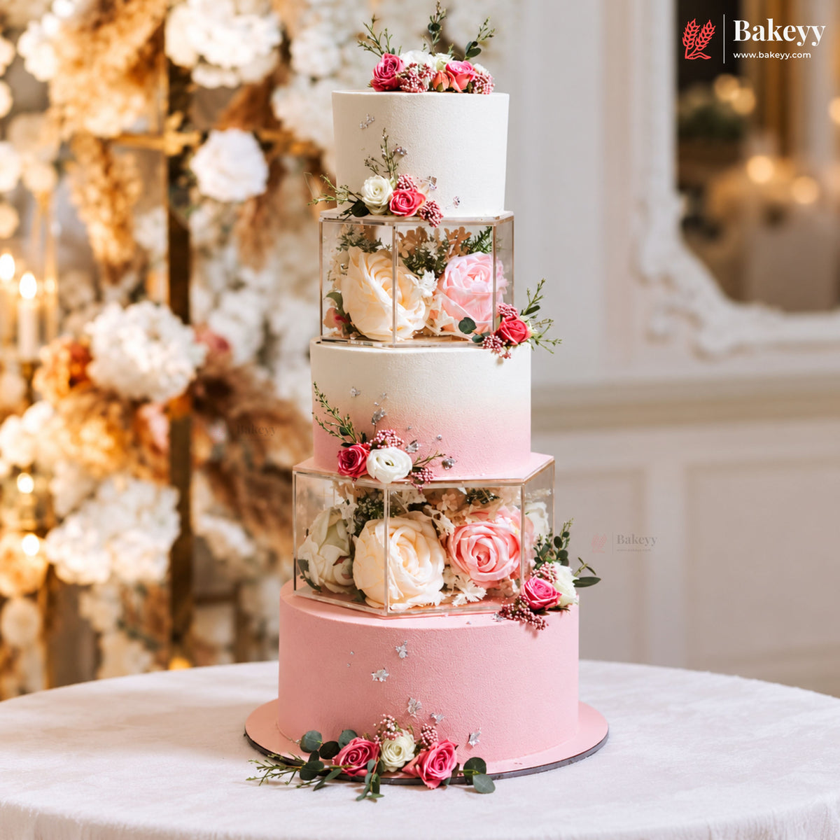 A five-tier pastel pink and white wedding cake decorated with pink and cream roses in clear square boxes, displayed on a white tablecloth with a blurred floral background. The Bakeey brand logo is visible in the top right corner.