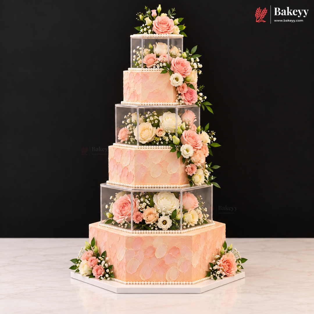 A five-tier peach and pink floral cake with clear acrylic cube risers, displayed on a white marble surface against a black background. The Bakeyy logo is visible in the top right corner.