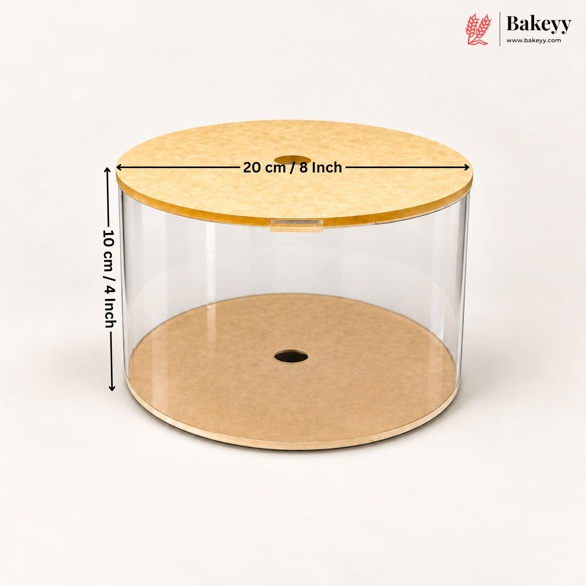 A clear cylindrical cake or pastry display cylinder with a light wood lid and a kraft paper base, shown on a white background with dimension arrows indicating 20 cm / 8 inch in diameter and 10 cm / 4 inch in height. Bakeyy logo is visible in the top right.