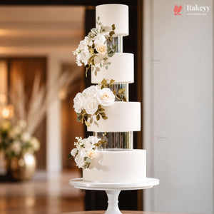 A five-tier white wedding cake with floral decorations and clear cylindrical inserts, displayed on a white pedestal stand in a softly lit indoor setting.