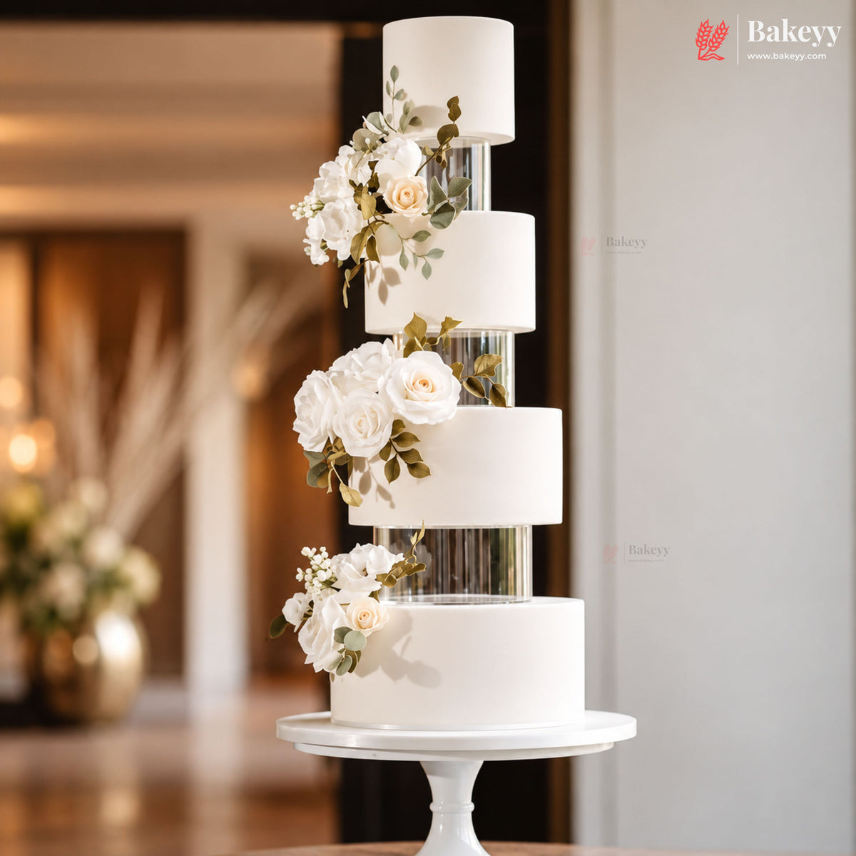 A five-tier white wedding cake with floral decorations and clear cylindrical inserts, displayed on a white pedestal stand in a softly lit indoor setting.