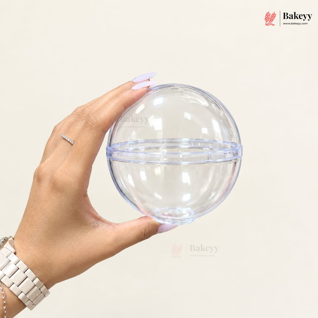 A hand holding a clear plastic round packaging dome with a horizontal seam, shown against a plain light beige background with the Bakeyy brand logo.
