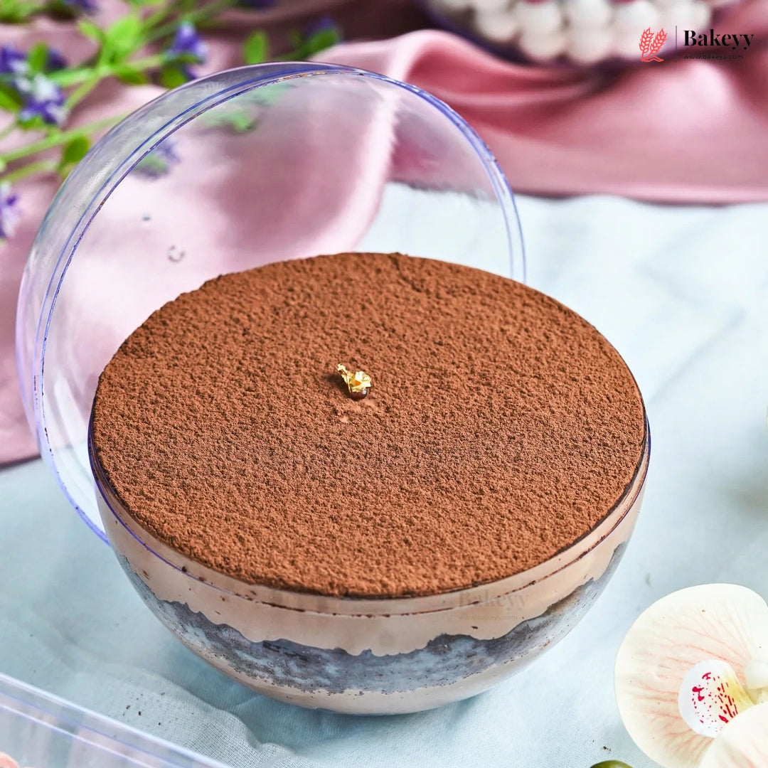 A layered dessert in a clear glass bowl, topped with a thick layer of cocoa powder and a small gold decorative piece, with the clear lid leaning behind it on a light blue surface.