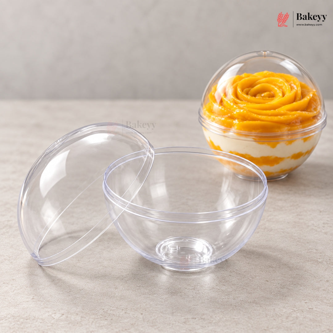 Two clear plastic dome dessert bowls on a light gray surface, with one bowl open and the other closed containing a layered yellow and white dessert. A Bakeyy logo appears in the top right corner.
