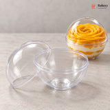Two clear plastic dome dessert bowls on a light gray surface, with one bowl open and the other closed containing a layered yellow and white dessert. A Bakeyy logo appears in the top right corner.
