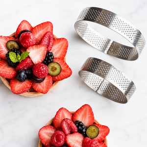 Two metal ring molds next to a bowl of sliced strawberries and berries on a white surface.