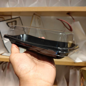 A hand holding a clear plastic clamshell container with a black tray insert, shown in front of a wooden shelf with white bags in the background.