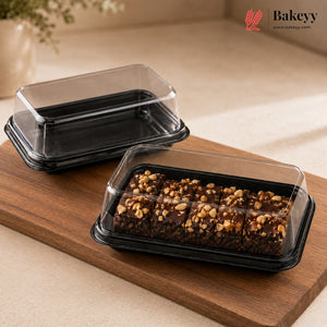 Two black plastic bakery boxes with clear lids, one empty and one holding chocolate bars topped with nuts, displayed on a wooden board with a Bakeyy logo in the corner.