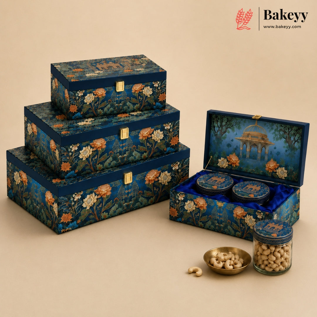 A set of ornate navy blue gift boxes with floral and architectural illustrations, shown closed in a stacked arrangement and open to reveal two round tins and a small bowl with nuts. A glass jar of nuts sits beside the boxes on a warm beige background.
