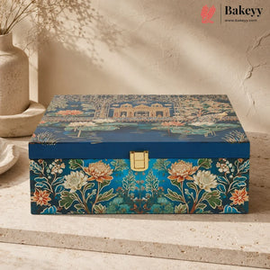 A decorative rectangular storage box with a blue lid featuring a garden pond scene and a floral front panel, shown on a stone surface against a textured beige wall. The Bakeyy logo appears in the top right corner.