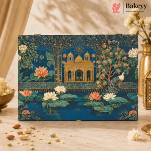 A rectangular decorative box with an ornate blue and gold illustration of a palace, peacock, lotus flowers, and trees, displayed on a beige surface with scattered nuts and flowers around it. The Bakeyy brand logo appears in the top right corner.