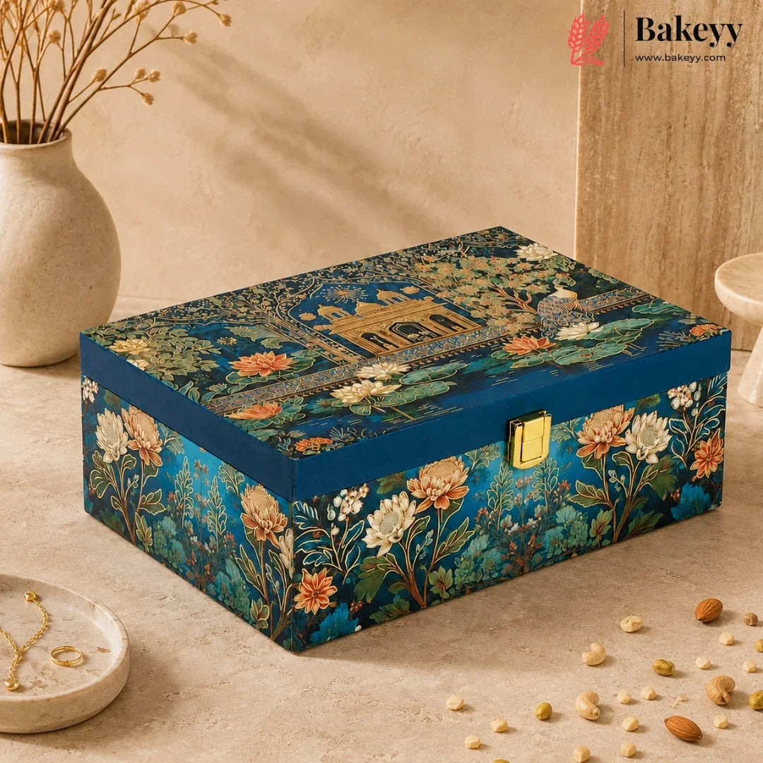 A rectangular decorative gift box by Bakeyy, featuring an intricate classical architectural and floral pattern in deep teal, gold, and orange. The box has a gold-tone metal clasp and is styled on a beige stone surface with a ceramic vase and scattered almonds nearby.
