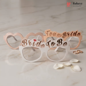 Bride to be glasses with 'Team Bride' text on a marble surface with rose gold rings and petals.