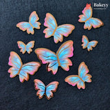 Seven butterfly-shaped sugar cookies with iridescent pastel wings in shades of blue, pink, and purple, arranged on a dark textured background. The Bakeyy logo is visible in the top right corner.