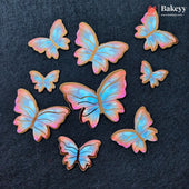 Seven butterfly-shaped sugar cookies with iridescent pastel wings in shades of blue, pink, and purple, arranged on a dark textured background. The Bakeyy logo is visible in the top right corner.