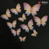 Nine butterfly-shaped cake toppers in pastel pink, purple, and blue with gold outlines, arranged on a dark textured background. The Bakeyy brand logo is visible in the top right corner.