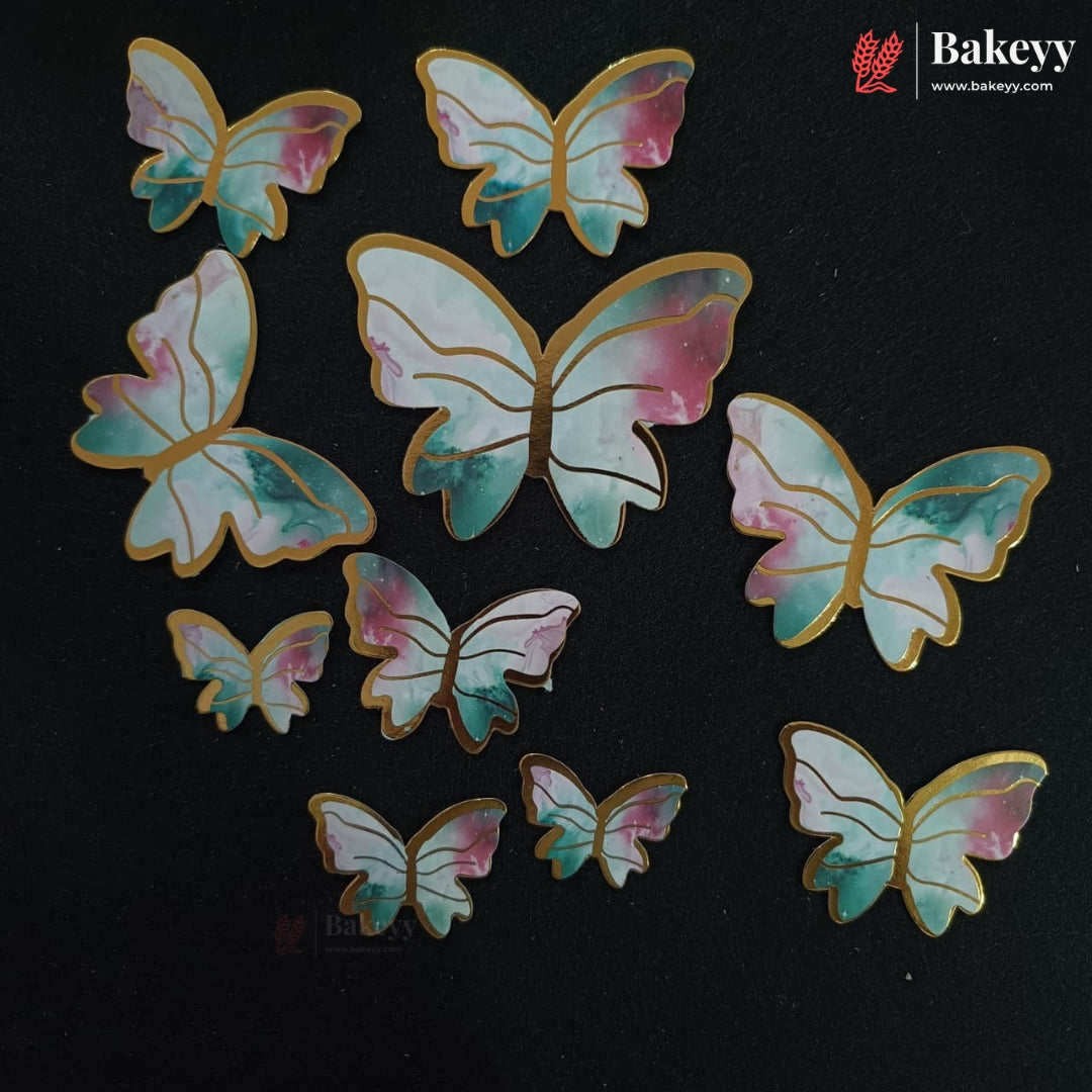 A set of butterfly-shaped decorative candies or stickers with a watercolor design in pastel pink, teal, and white, outlined with gold foil edges, arranged on a black background. The brand name 'Bakeyy' is visible in the top right corner.