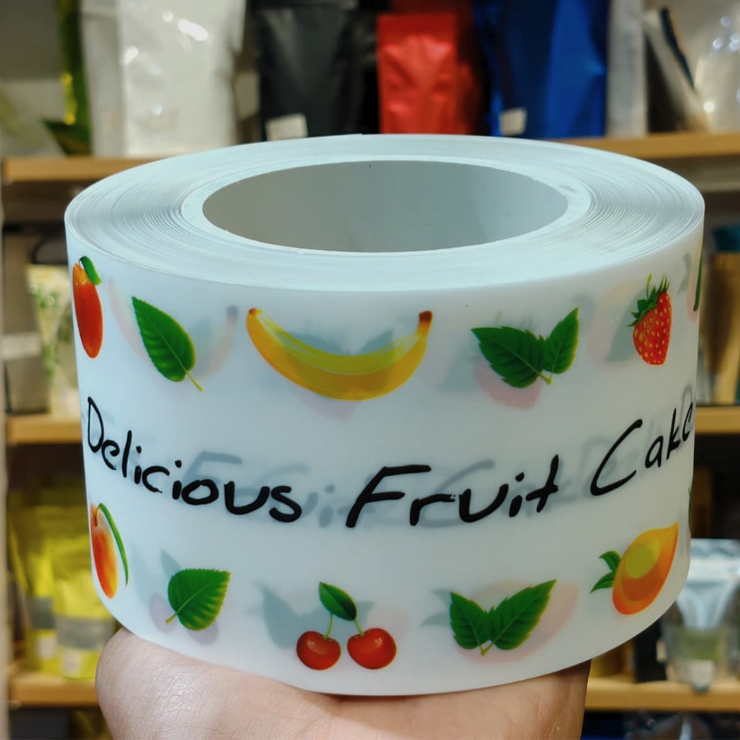 A hand holds a roll of white decorative labels printed with illustrations of fruit and the text 'Delicious Fruit Cake' in black script, with a blurred store background.