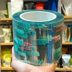 A hand holds a roll of clear decorative tape featuring a botanical pattern of teal leaves and small pink flowers, with a blurred shop background.