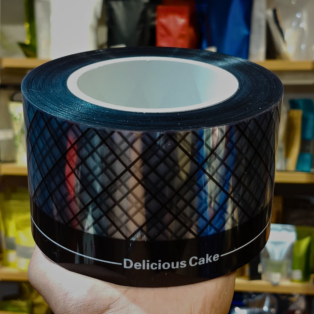 A hand holds a roll of black tape printed with a diamond grid pattern and the text 'Delicious Cake' on a blurred store shelf background.