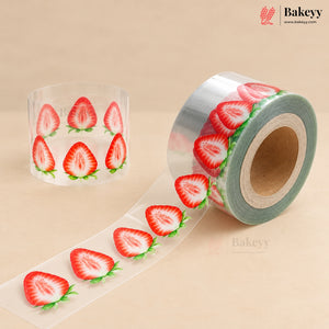 A roll of clear adhesive tape printed with red strawberry slices and green leaves, shown unspooled alongside a standing spool on a beige background. The Bakeyy brand logo is visible in the top right corner.