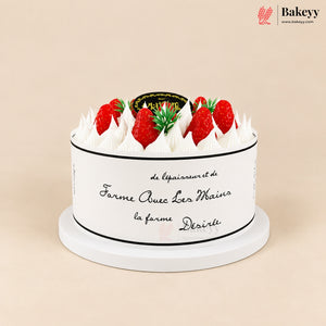 A round white cake with piped white frosting on top, decorated with fresh red strawberries and green leaves. The cake has black cursive French text on the side and a small black and gold decorative topper on top.