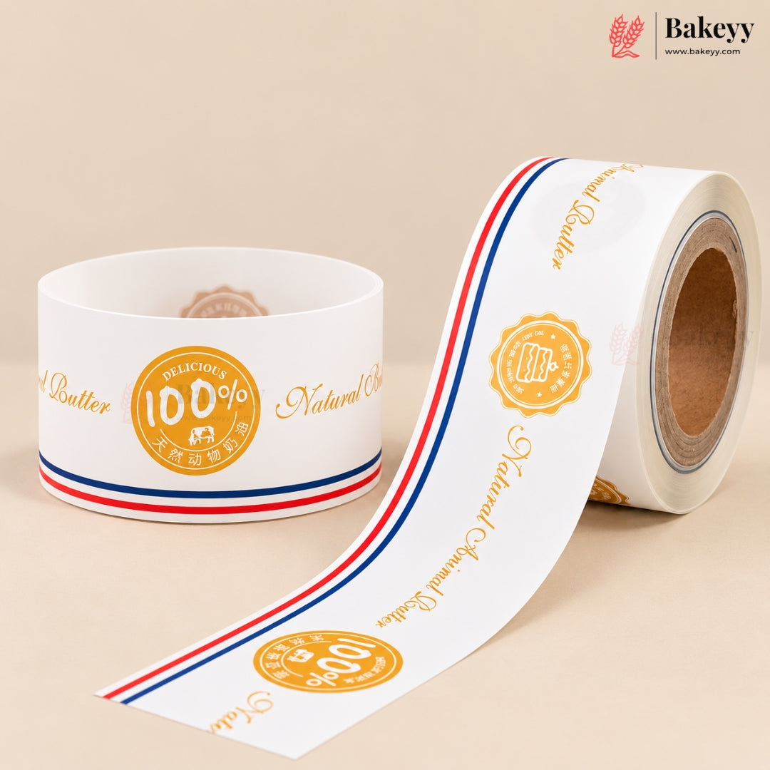 A roll of white bakery label tape with gold text and a red, white, and blue stripe, shown next to a spool on a beige background. The brand 'Bakeyy' is visible in the top right corner.