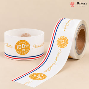 A roll of white bakery label tape with gold text and a red, white, and blue stripe, shown next to a spool on a beige background. The brand 'Bakeyy' is visible in the top right corner.