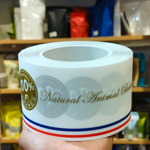 A hand holds a roll of white paper labels printed with 'Natural Animal Based' in gold script, a gold circular '100% Natural' seal, and red and blue stripes at the bottom, with a blurred store shelf in the background.