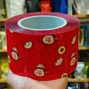 A hand holding a roll of red tape printed with Chinese New Year motifs, including the character 福, gold ingots, and copper coins, against a blurred background of shelves.